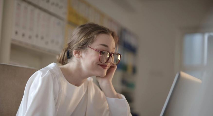 Woman in naturally-lit room with glasses reading on a laptop