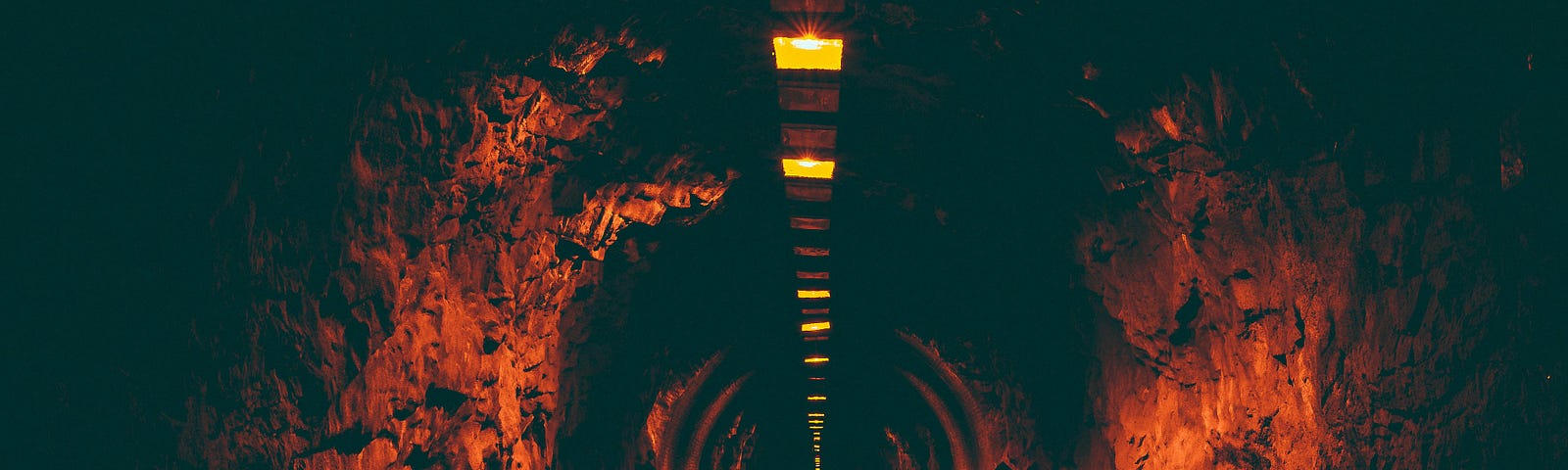A dark tunnel with overhead lights turned on. The orange glow of the lights reflects on the wet road.