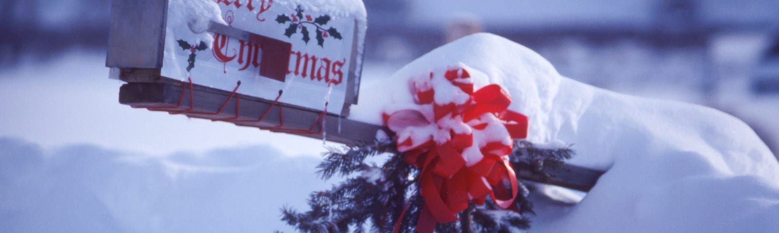 Mailbox with the words “Merry Christmas,” a red bow, and a small evergreen tree branch protrude from a snow bank.