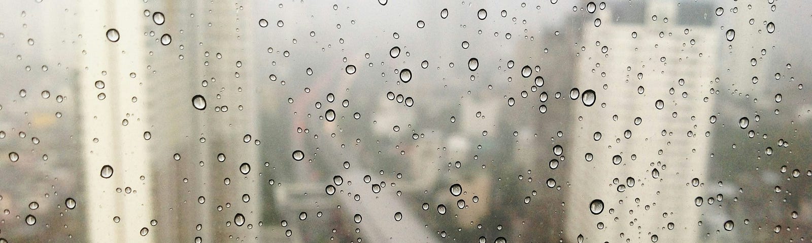 A city view from a window high up: the window is covered in rain drops, and the sky is a silvery grey.