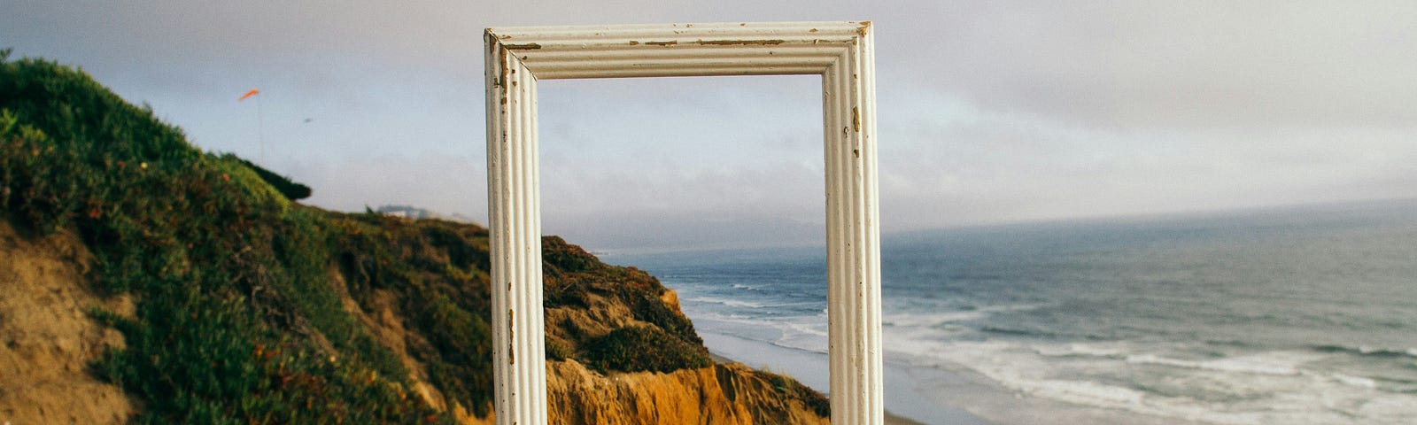 A hand holding up an empty white, wooden frame against the seaside landscape.