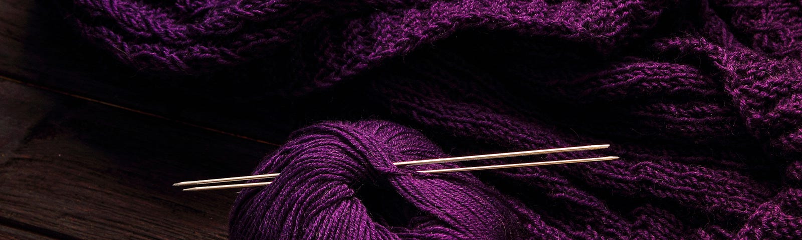 A purple ball of wool some knitting, and knitting needles pushed through the ball.