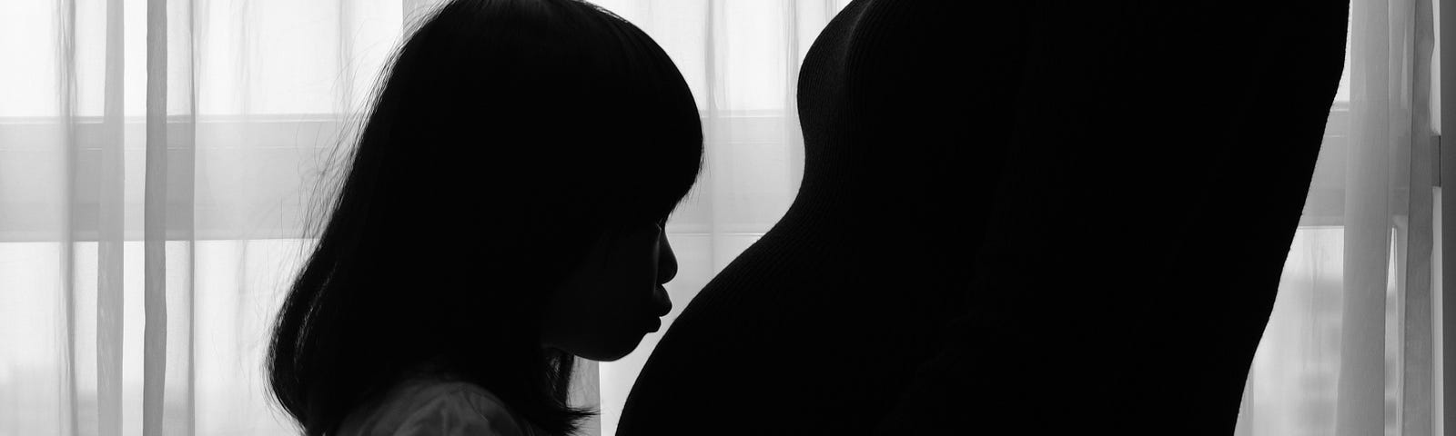 Black and white photo of a preschool girl kissing her mom’s pregnant belly.