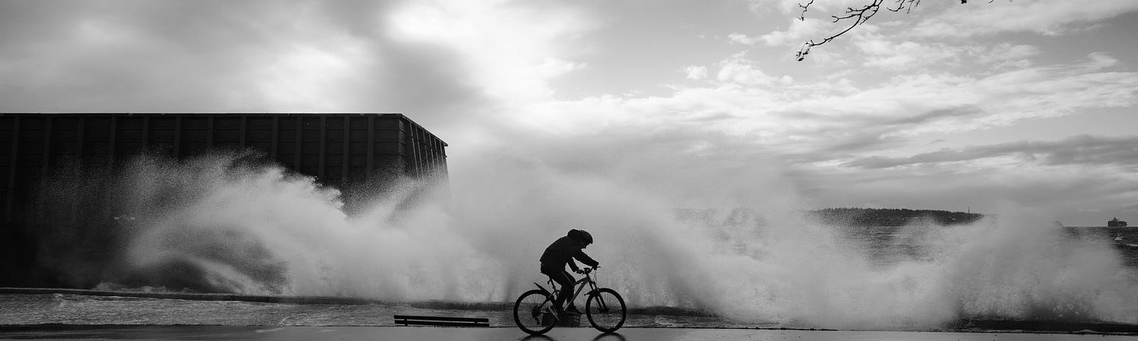 A cyclist rides along a wet road as waves crash over the sea wall behind, and the sun breaks through the clouds