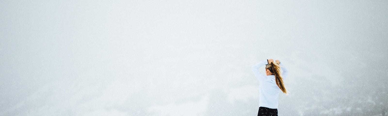 Young woman in black pants and white jacket standing at the precipice of a white glacier.