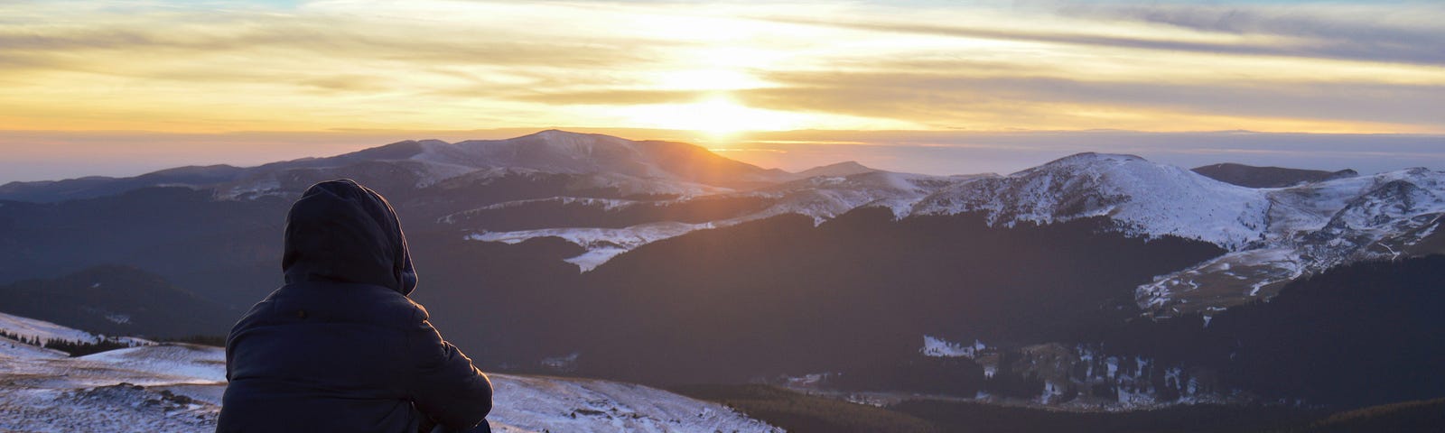 Human wearing jacket sitting mountainside, knees bent, back to the camera, watching the sun rise over snowy mountains.