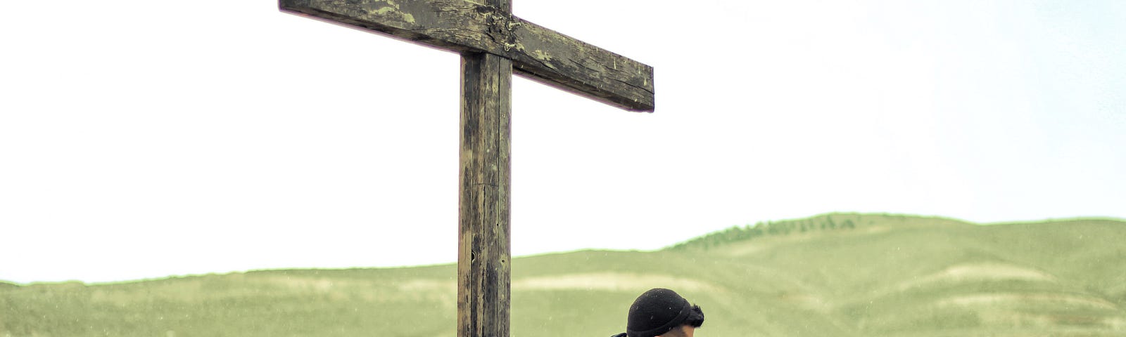 A man sitting at the foot of a cross with the green mountains behind him.