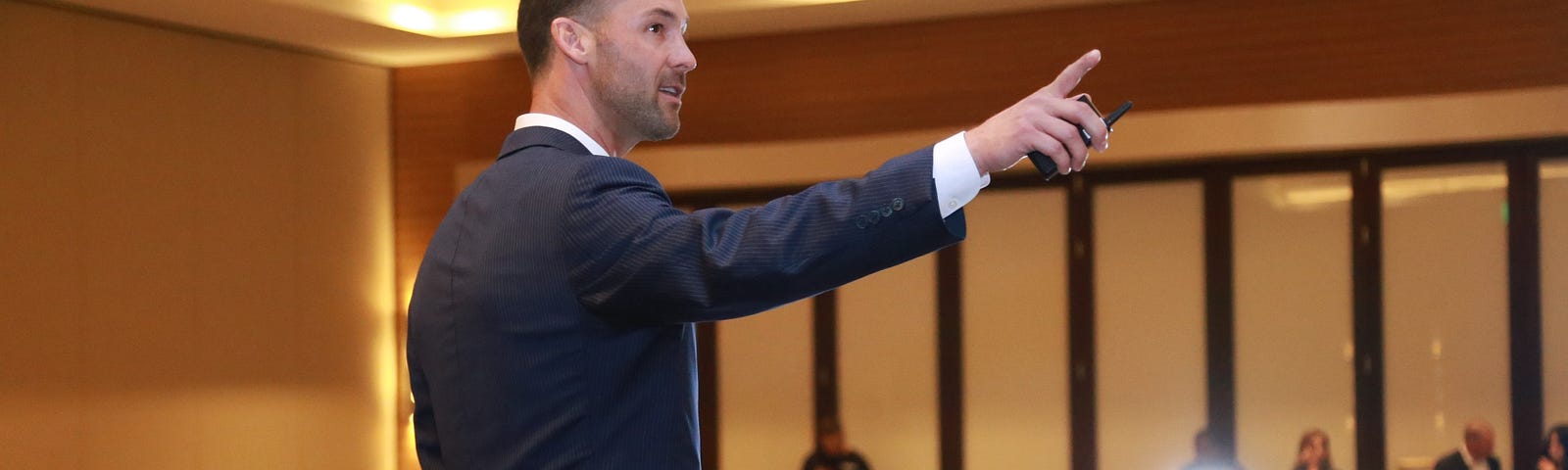 A white man in a suit gestures to something off camera for an audience