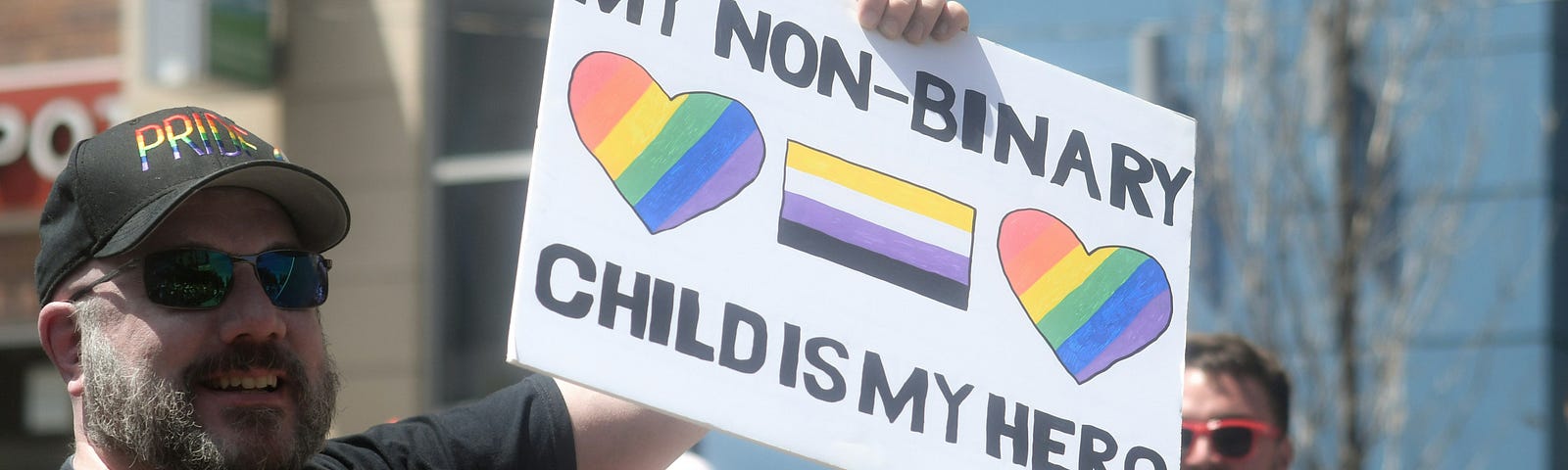 A man at a pride parade wears pride hat and t-shirt. He’s holding up a sign that says, “My non-binary child is my hero” and it has the pride and trans flag on it.