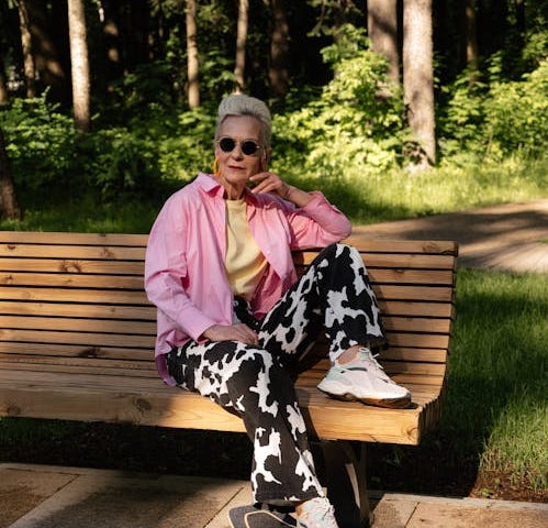 Elderly white woman with short white hair, big orange hoop earrings, and black sunglasses, wearing a pink button-down shirt with a yellow top underneath, with velvet black-and-white jersey cow patterned pants and white sneakers. She’s sitting on a wooden bench with one foot on a black skateboard, in partial sun at a park.