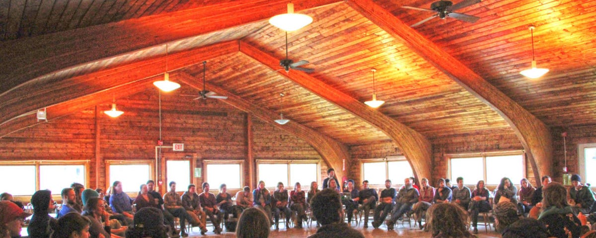 A community gathering of people sitting in a semi-circle in a large space with a high arched wooden-plank ceiling.