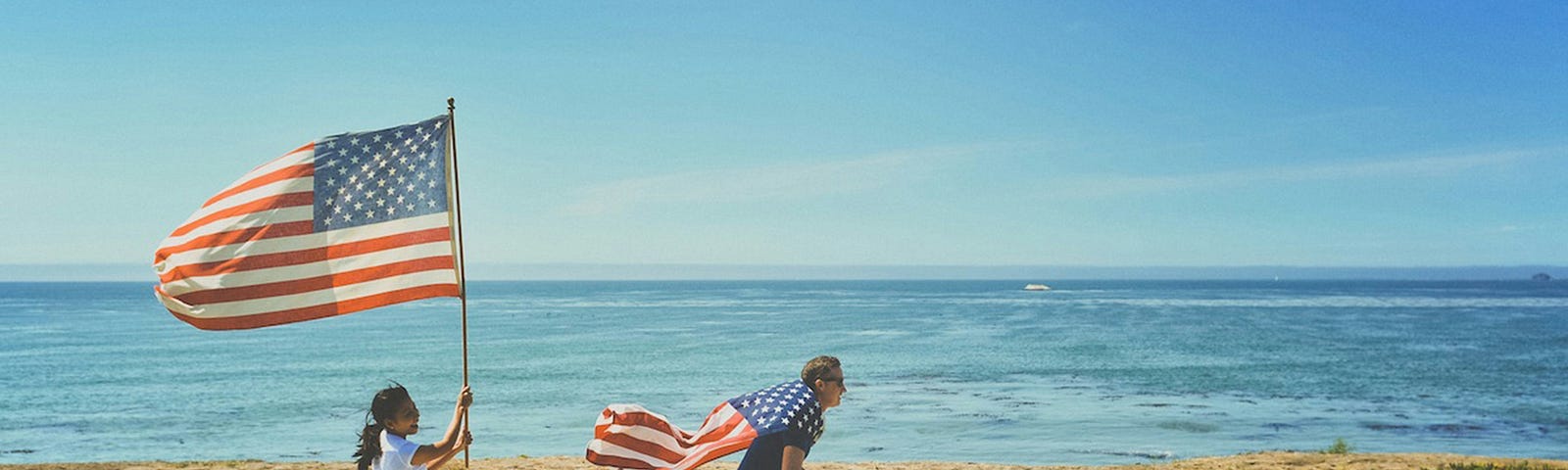 A man riding a bike along the beach, with an American flag tied around in neck, fluttering behind. A young girl, running behind him with a big American flag behind him.