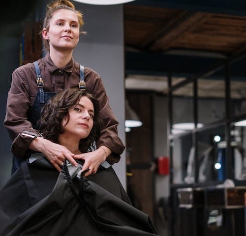 A hairstylist is securing a protective plastic cape over a young woman’s shoulders in preparation for a haircut.