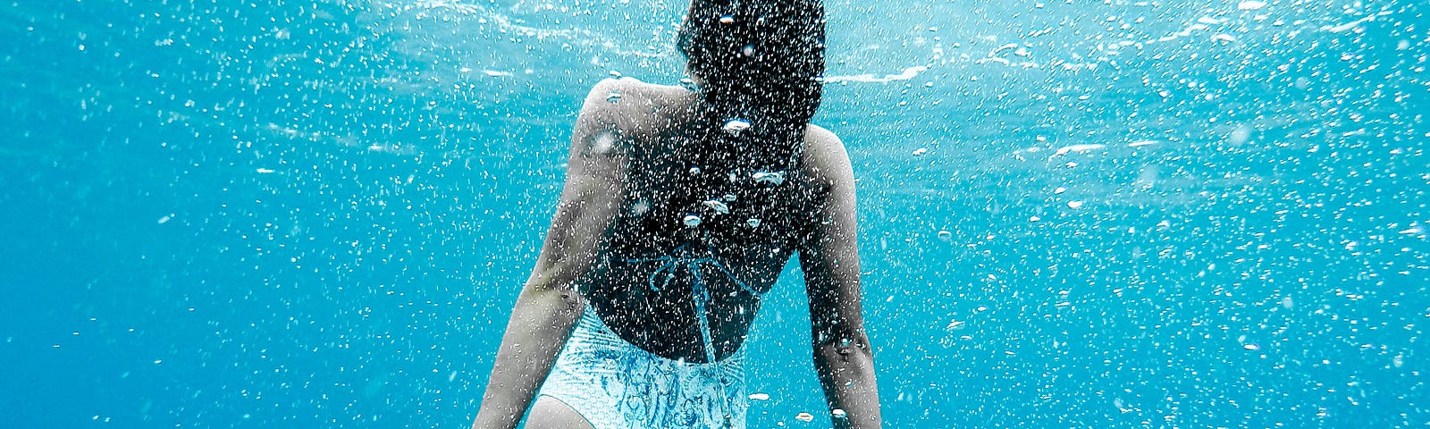 A Woman in a white bathing suit, as seen from behind. She swims towards the surface of the open water, her arms pointed down at at a 30 degree angle from her body. The water is light blue and their are bubbles within it.