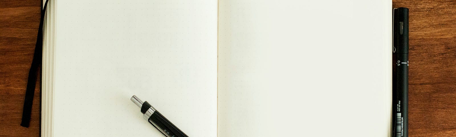 A photograph of an open notebook with a small area of the desk it’s sat on in view around the edge. A pen lies across the notebook.