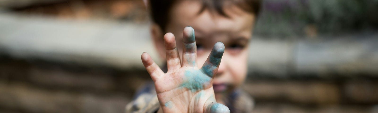Portrait of toddler boy outside in front of a stone wall with stained blue hand reaching out towards the camera. The hand is in focus, the child’s face is out of focus and partially blocked by his hand.