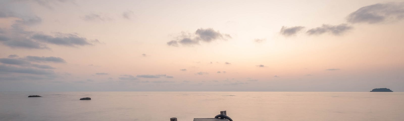 Pale grey-blue sky with scattered clouds, above a grey-blue ocean, with a wooden dock protruding out over the water in the foreground. On the horizon is a beautiful peachy-pink sunrise or sunset.