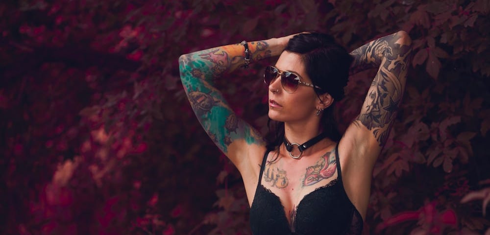 Standing woman wearing sunglasses with bright tattoos on her arms torso and upper chest.
