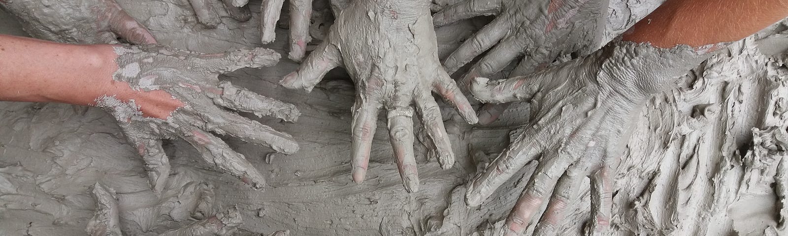 Nine small hands covered in modeling clay, fingers extended in a circle towards each other over a board covered in the same gray clay.