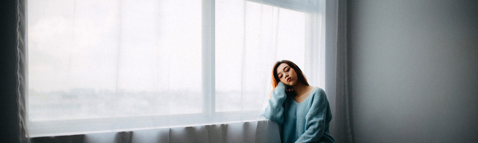 A woman, wearing a blue jumper and orange socks, sits on the floor in an empty room. She is leaning her elbow on a window sill. White curtains hang from the wide window. She looks sad.