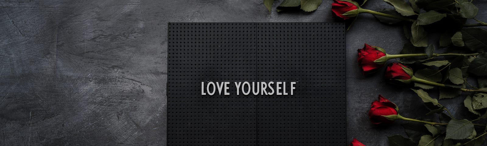 A black background with white block letters reading, “LOVE YOURSELF.” On the right, several red roses lie pointing left.