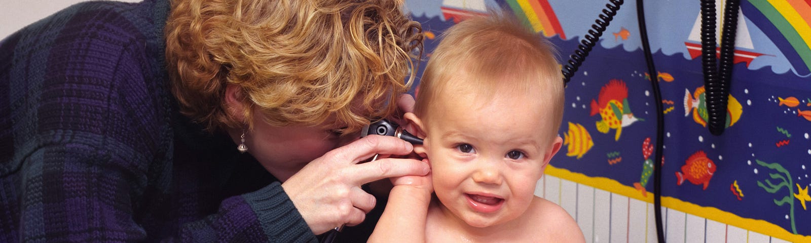 A baby sitting on an examination table is being checked by a healthcare professional using an otoscope to look into the baby’s ear. The background is decorated with a colorful wallpaper featuring rainbows and aquatic life.