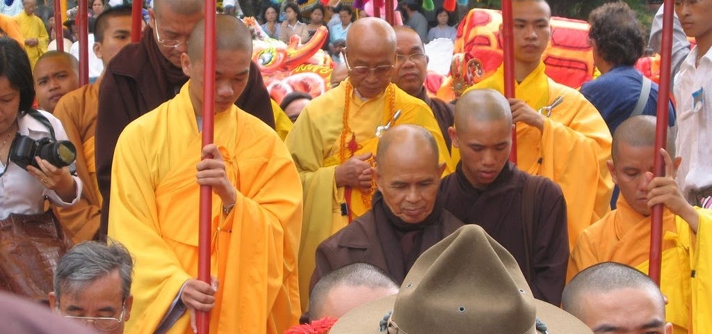 Thich Nhat Hanh in brown Buddhist robes surrounded by other Buddhist monks in yellow robes.