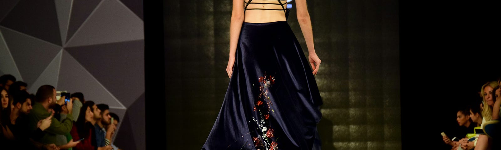View from the back of a model on a catwalk wearing a long black gown with thin straps crossed across the back.