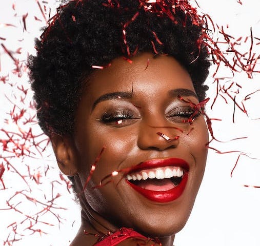 Head and shoulder shot of smiling woman in red strap dress being covered in tinsel confetti who looks happy and like she is celebrating