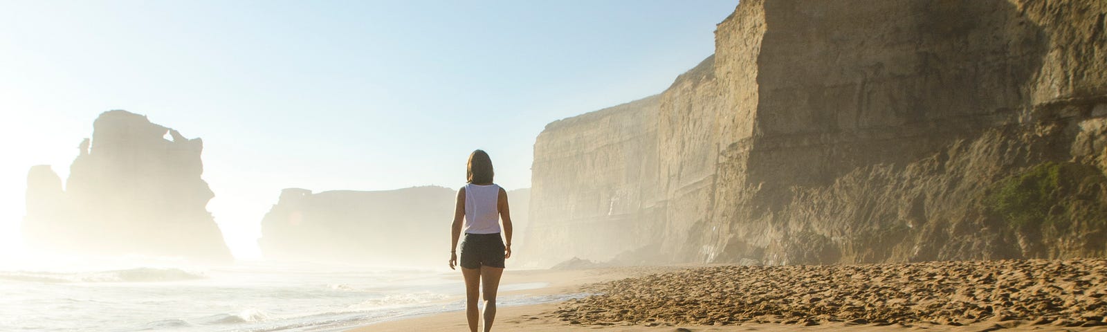 A woman walks away from us on a sandy beach. Cliffs loom in the background. Physical activity is associated with lower colorectal cancer risk.