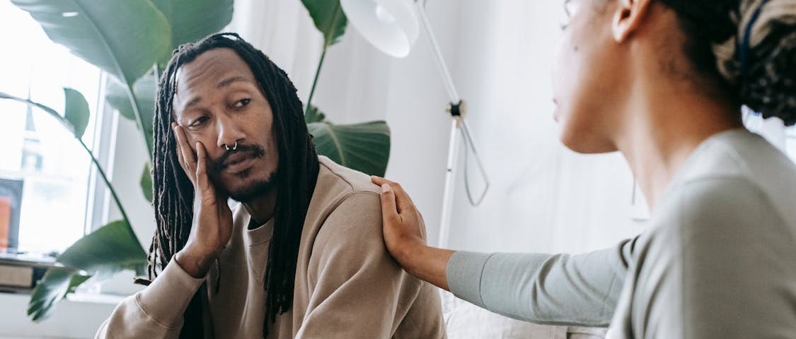 A concerned woman gently places her hand on a man’s shoulder as they have a serious conversation in a cozy, plant-filled room.