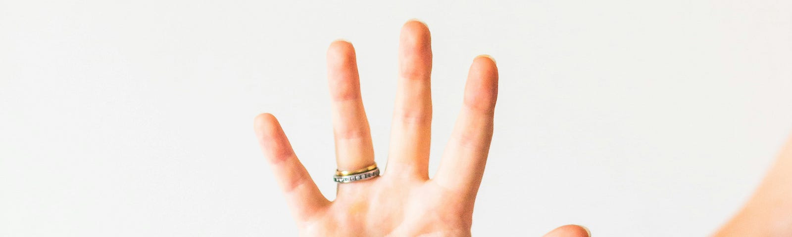A hand is held up with all fingers spread, as if to say “five” or “that’s enough”. The person wears two thin bands on the fourth finger; one studded with diamonds, the other gold.