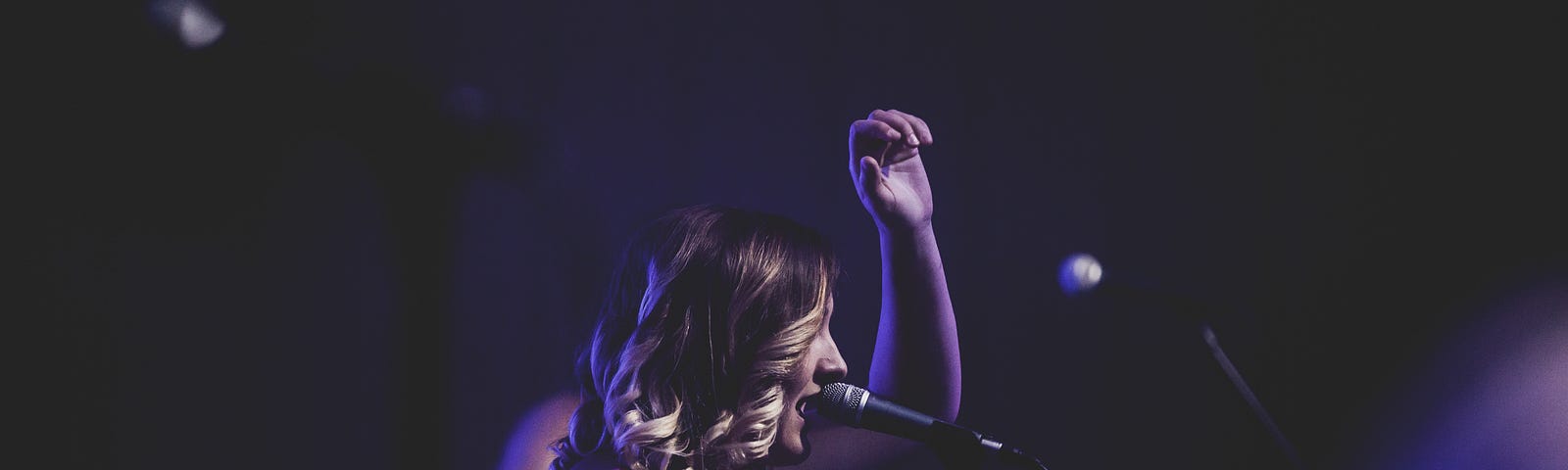 A dark background with a woman in purple in profile lifting one hand in worship while she sings into a mic on a stand.