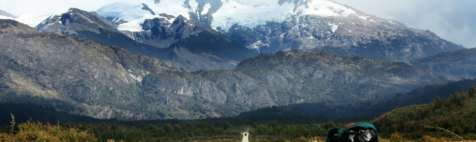 A landscape showing a gravel road through shrubland, with a snow-capped mountain on the horizon. A bike loaded with traveling gear is in the foreground, though the rider is nowhere to be seen.