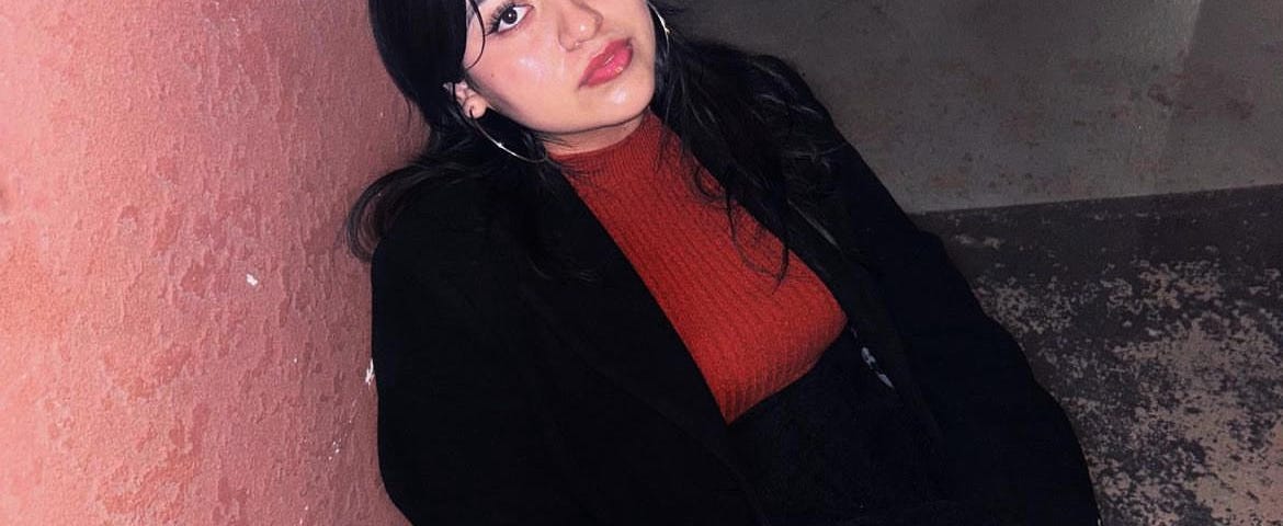 Shelby Silva is sitting outside and leans against a pink wall. She has her legs crossed and she’s wearing a black blazer over a red knit sweater. She wears a black skirt and sheer leggings. There is an iron table next to her.