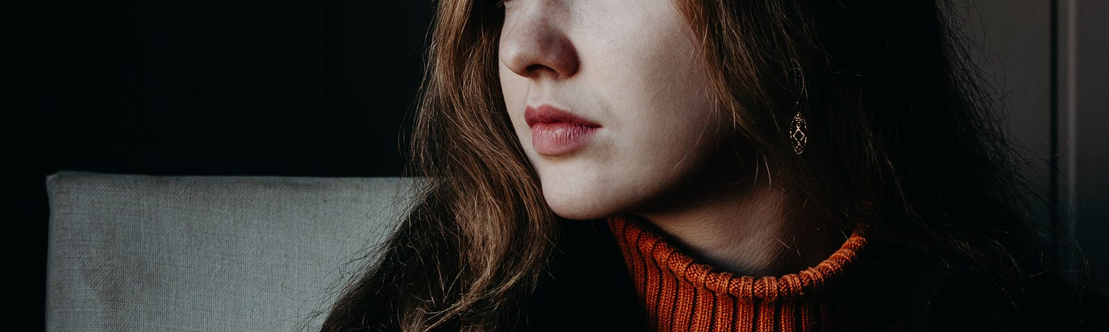 A woman with long brown hair and wearing a jumper and jacket, gazes out a window. She is holding a black phone and a coffee cup sits on the table in front of her.