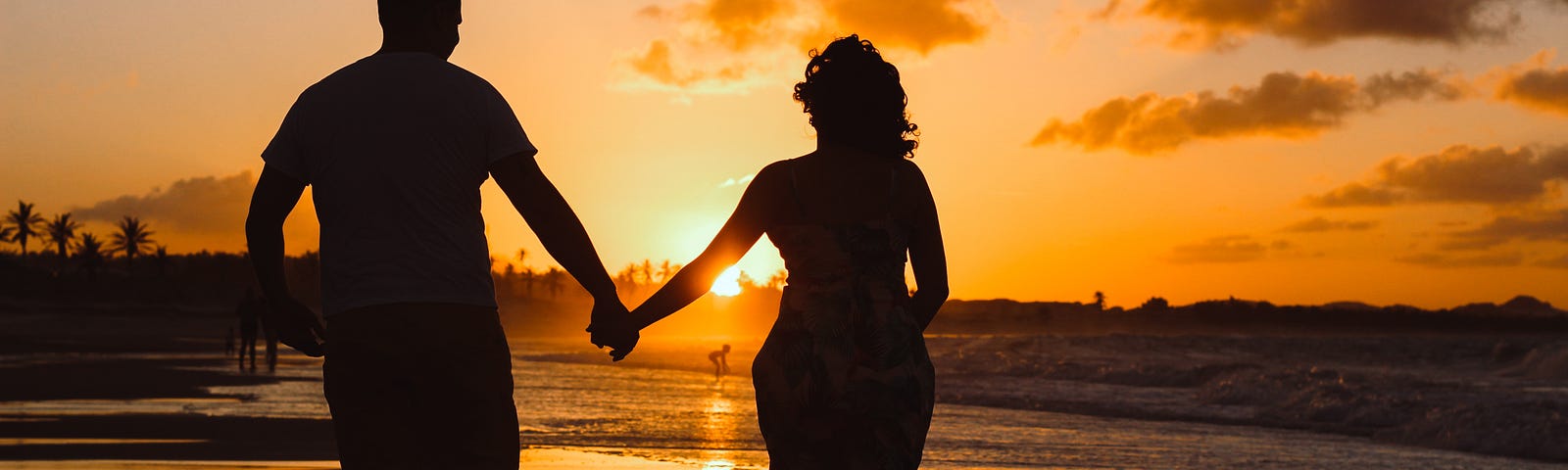 A couple holding hands and walking on the shore with a sunset behind them.