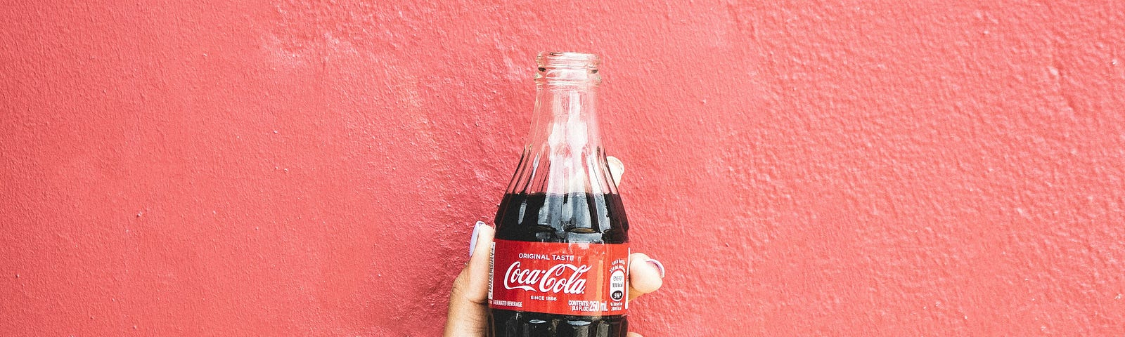 A hand holds a bottle of cola in a glass bottle against a red painted wall.