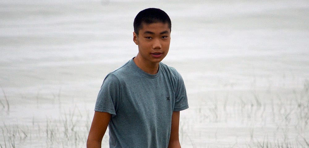 Photo of a teenaged boy wearing a t-shirt, standing in front of a body of water.