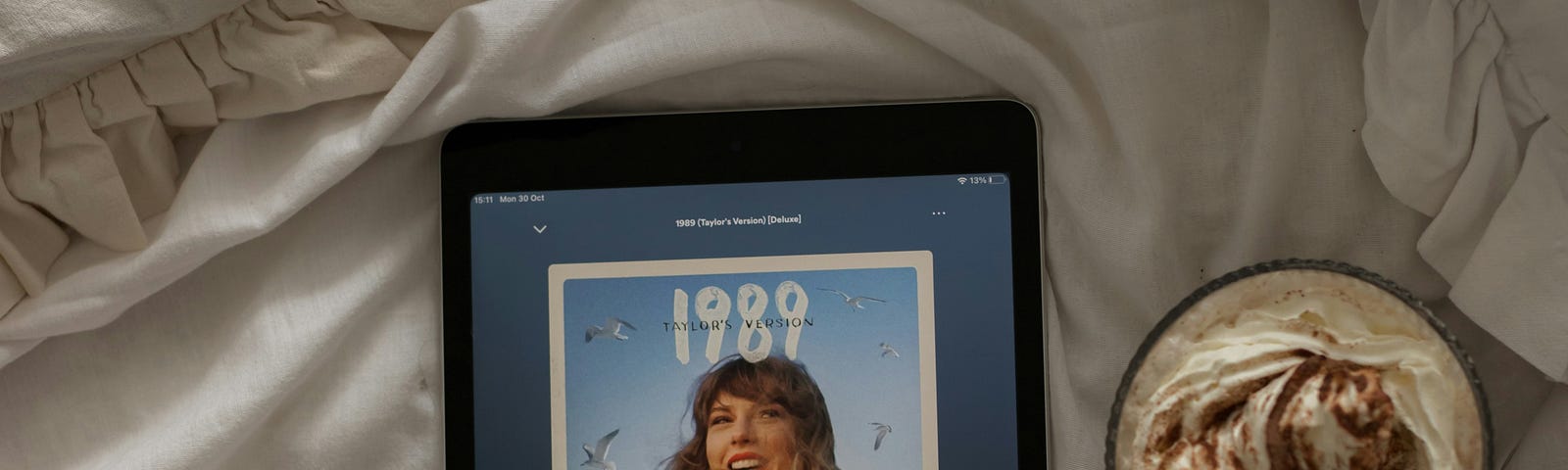 An iPad screen showing a screenshot of Taylor Swift, the iPad is on a bed with crumpled cream sheets, and there is a cup of cappuccino next to the iPad