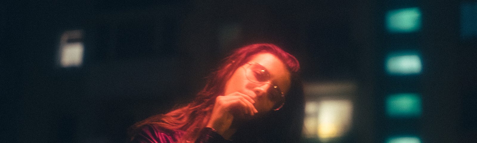 Image with a black background and a woman with red hair, glasses, and a leather jacket cocking her head to the side and looking into the camera, her hand in front of her mouth.