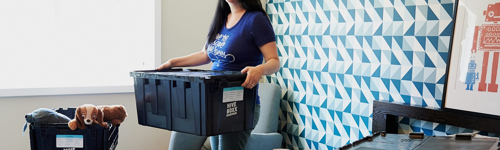 A woman and small child appear to be packing up the child’s bedroom. There are boxes with toys around the room. The woman is carrying one of them and smiling at something or someone outside of our view.