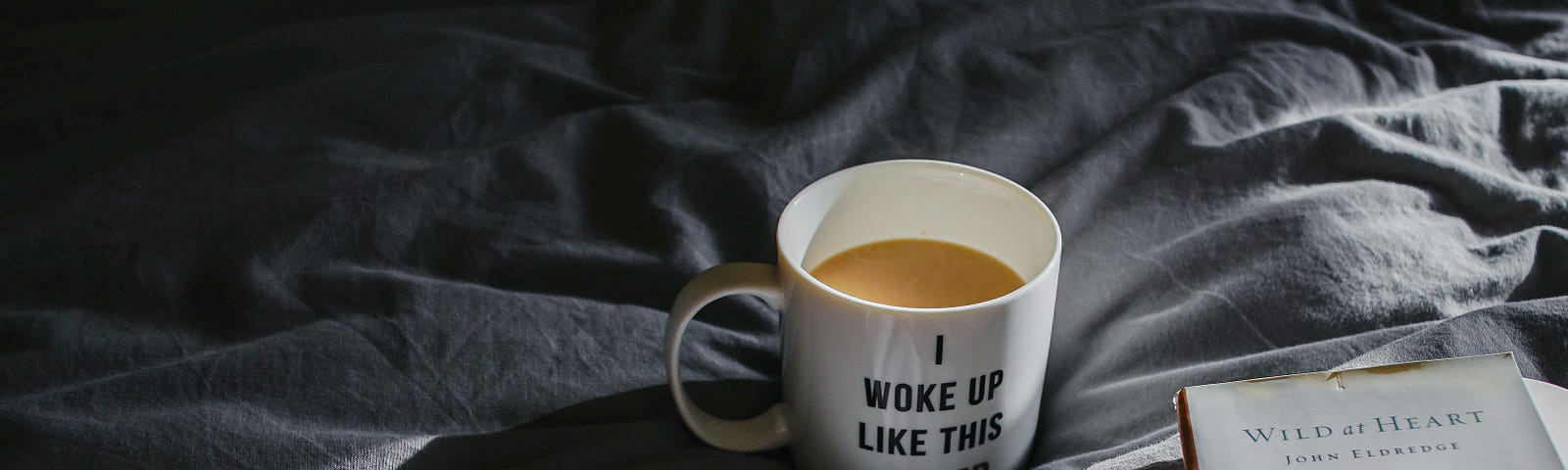 photo of a coffee mug that reads “I woke up like this #tired.” The mug is sitting on gray sheets beside an open book.
