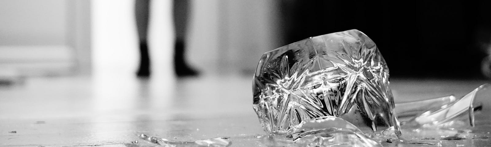 Black and white picture of a broken decrorative glass in the foreground and the legs of a person blurred in the background