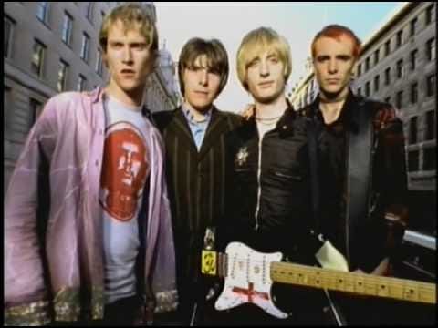 From Kula Shaker “Hey Dude” music video; four band members with frontman Crispian wearing his black&white stratocaster guitar with a city backdrop