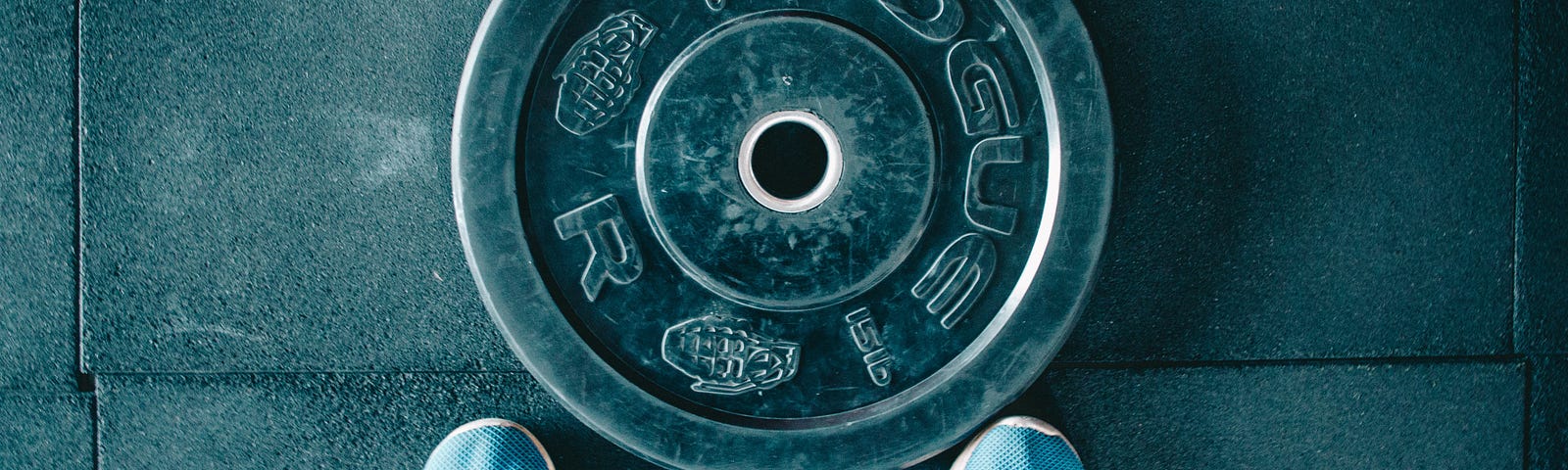 A 15-pound weigt plate sits on the floor in front of a person’s two blue shoes. We see the scene from above. Testosterone is crucial in maintaining my vitality, energy levels, and overall well-being.