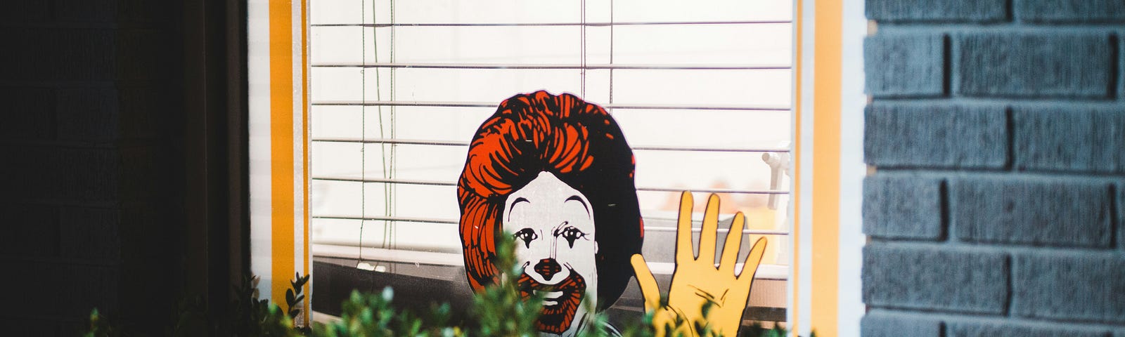 A cutout of Ronald McDonald peers out from against the wall of a house, behind a bush.