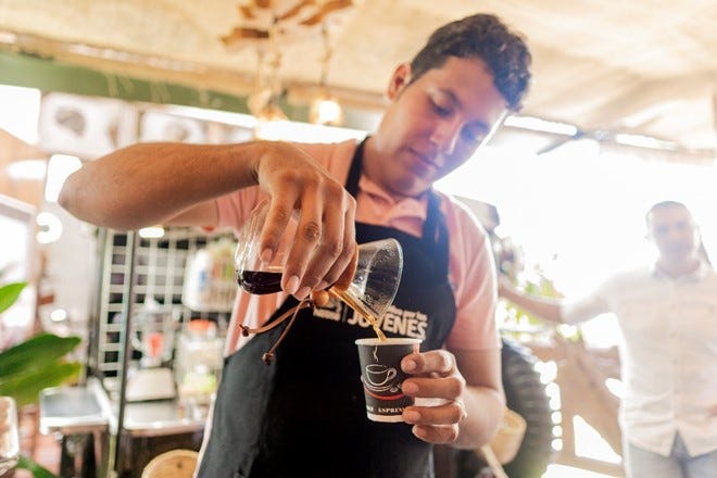 A young man wearing an apron pours coffee into a cup.