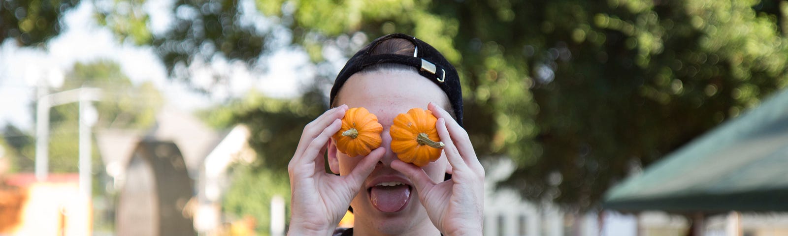 A person holds small pumpkins to their eyes and sticks out their tongue
