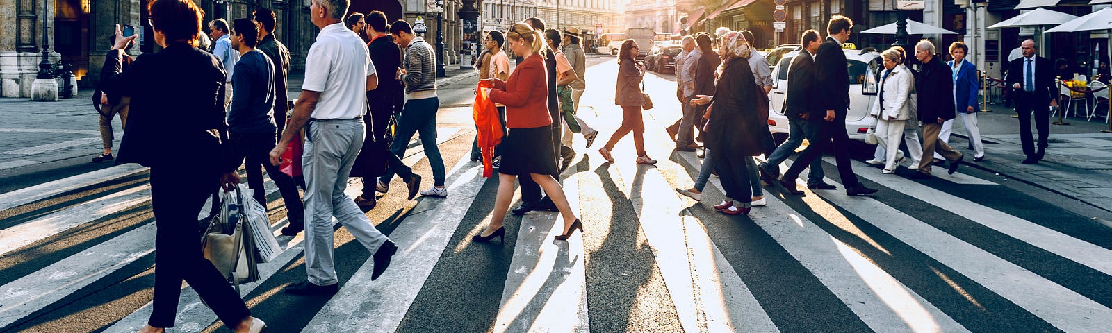 A busy street walking intersection filled with people. The Sun is rising or setting in the distance. City sprawls hide the nuances of the sun’s setting or rising; yet the abundance of people crossing the street remains.